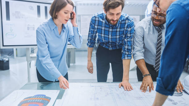 istock In the Industrial Engineering Facility: Diverse Group of Engineers and Technicians on a Meeting Gather Around Table with Engine Design Technical Drafts, Have Discussion, Analyse Technology 1176843575