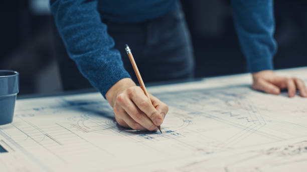 istock In the Dark Industrial Design Engineering Facility: Male Engineer Works with Blueprints Laying on a Table, Uses Pencil, Ruler and Digital Tablet. On Desktop Multiple Drawings. Focus on Hands 1176843605