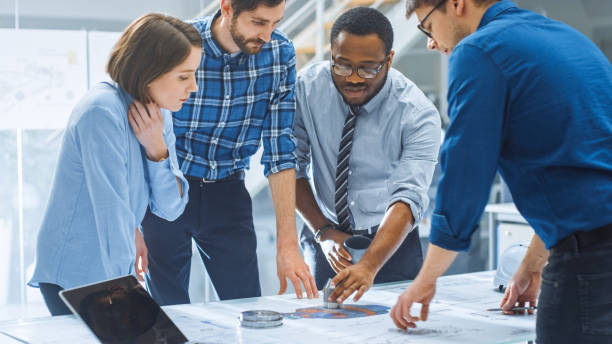 istock In the Industrial Engineering Facility: Diverse Group of Engineers, Technicians, and Specialists on a Meeting, Have Discussion, Analyse Engine Design Technical Drafts that are Lying on the Table 1176843628