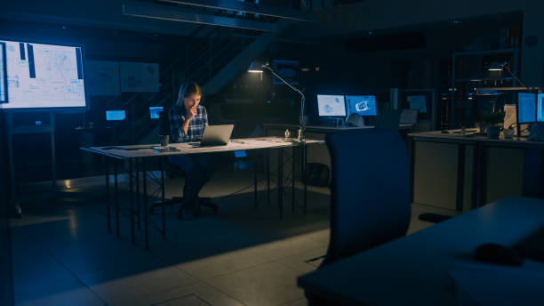 istock Beautiful Female Engineer Sitting at Her Desk Works on a Laptop Computer. Blueprints Lying on a Table. In the Dark Industrial Design Engineering Facility. Warm Light Lamp 1176843753