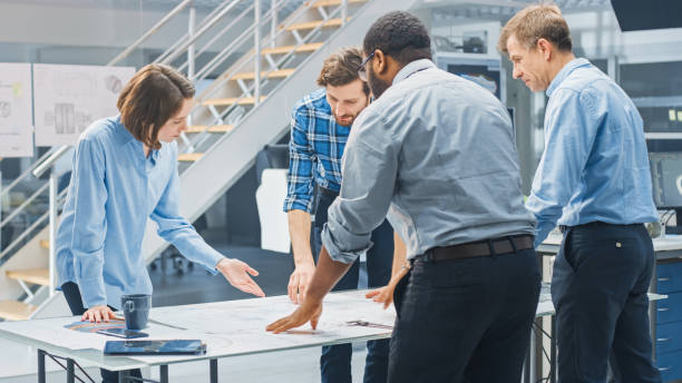 In the Industrial Engineering Facility: Diverse Group of Engineers and Technicians on a Meeting Gather Around Table Unravel Sheets of Engine Design Technical Drafts, Have Discussion, Analyse Drawings.