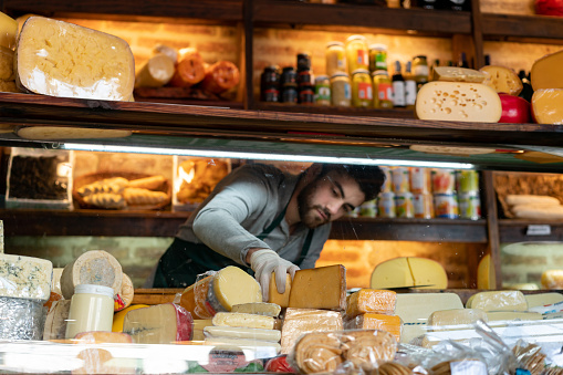 Young latin american man arranging the cheese refrigerator display at a delicatessen - Small business concepts
