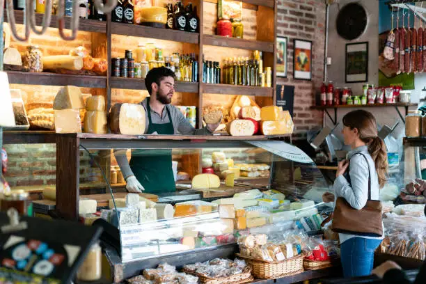 Photo of Latin american man working at a delicatessen suggeting a type of cheese to female customer