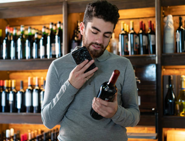 guapo cliente latinoamericano leyendo la etiqueta en una botella de vino mientras habla en un teléfono inteligente en una bodega - wine cellar audio fotografías e imágenes de stock