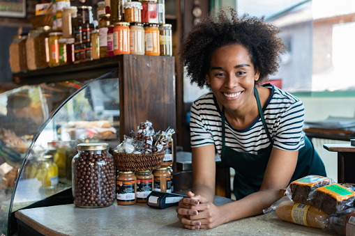 Friendly black saleswoman at a delicatessen leaning on counter smiling at camera very happy