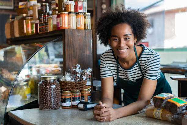 Friendly black saleswoman at a delicatessen leaning on counter smiling at camera Friendly black saleswoman at a delicatessen leaning on counter smiling at camera very happy convenience store photos stock pictures, royalty-free photos & images