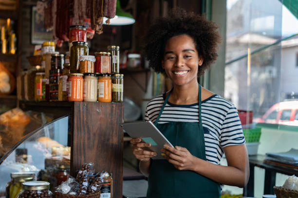 istock Cheerful black saleswoman at a delicatessen holding a tablet while facing camera smiling 1176845371