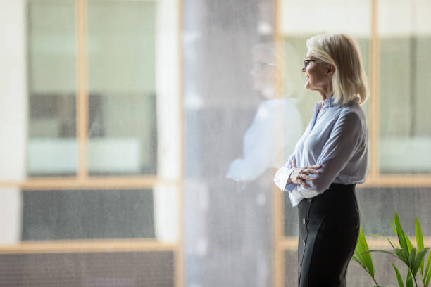 fiduciosa donna d'affari matura che riflette strategia, in piedi vicino alla finestra - thinking women businesswoman business foto e immagini stock
