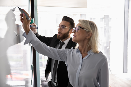 Mature businesswoman wearing glasses training employee, drawing on flip chart, mentor coach explaining business strategy, discussing marketing plan, colleagues working on project together