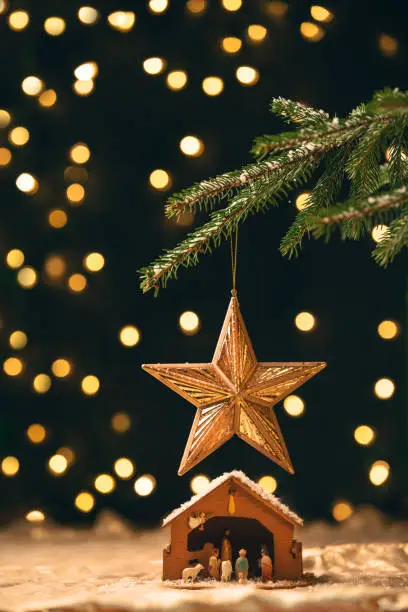 A manger sitting under a star ornament that is hanging from a Christmas tree with lights blurred behind.