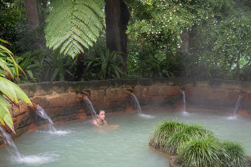 Furnas, Azores, Portugal -  August 10, 2019: Teen girl taking massage in a mineral thermal water hot tub pool, surrounded by lush green foliage in the Terra Nostra park at Furnas, Sao Miguel island, Azores.