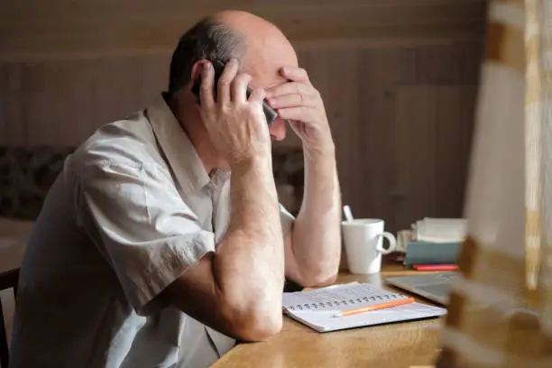 Senior man feeling upset having phone conversation depressed by hearing bad news sitting at home