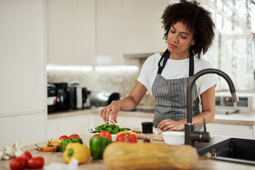 Attractive mixed race woman in apron standing in kitchen and taking rocket from plate. On kitchen counter are tomatoes, peppers and zucchini.