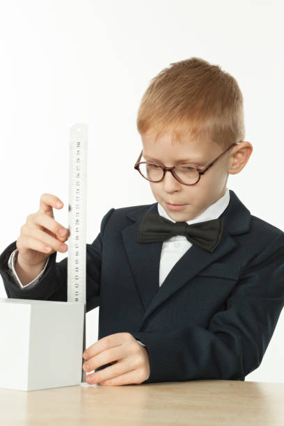 bambino di 7 anni in uniforme scolastica e occhiali misura le dimensioni di un cubo con un righello - little boys measuring expressing positivity intelligence foto e immagini stock