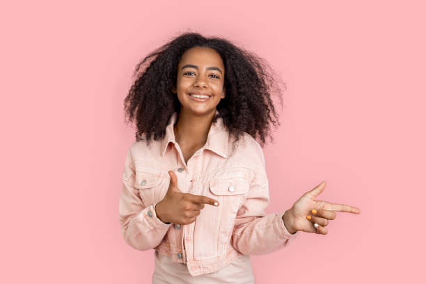 kür. afrikanische mädchen in jeansjacke stehen isoliert auf rosa zeigt beiseite auf kopierraum lächelnd fröhlich - afro amerikanischer herkunft fotos stock-fotos und bilder