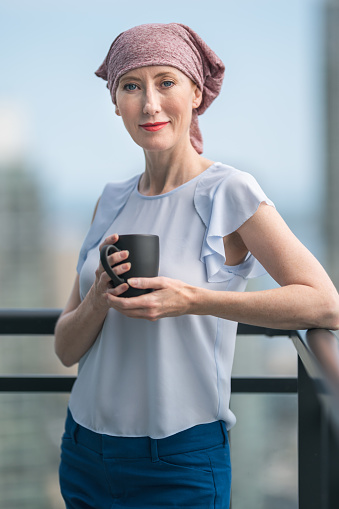 A caucasian woman with cancer is wearing a bandana to hide her hair loss. She is standing on a patio of a skyscraper. She is holding a cup of tea while admiring the urban skyline. The woman is smiling directly at the camera. She is filled with gratitude and hope for recovery.