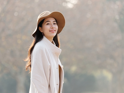 Portrait of beautiful Chinese young woman in beige coat and hat posing and smiling at camera, charming Chinese girl with black long hair enjoy her leisure time outdoor.
