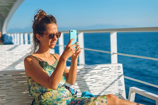 Young woman on the bench on deck of the ferry boat or ship yacht taking photos pictures using her smart mobile phone while sailing on the vacation in sunny day