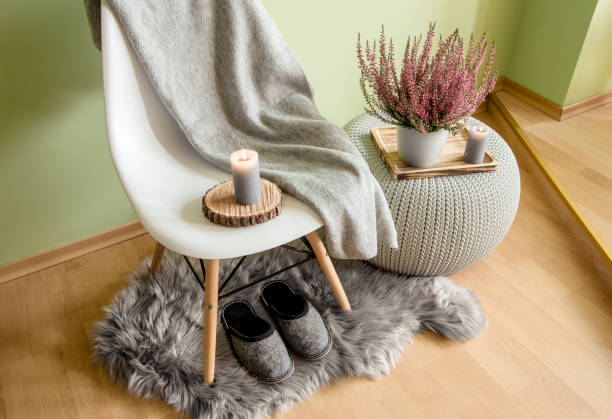 istock Plastic chair with wood legs covered with gray wool plaid, knitted pattern plastic table, fake sheepskin rug with felt slippers and heather in flower pot, against pale green wall. 1176967487