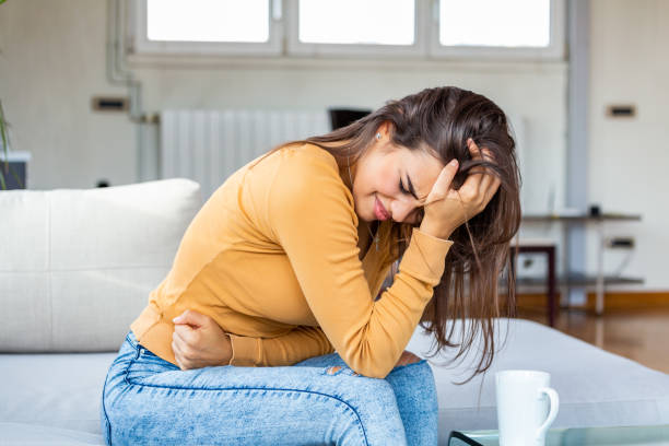 junge kranke frau mit händen halten drücken ihren schritt unterbauch. medizinische oder gynäkologische probleme, gesundheitskonzept. junge frau leidet unter bauchschmerzen beim sitzen auf dem sofa zu hause - eileiterschwangerschaft stock-fotos und bilder