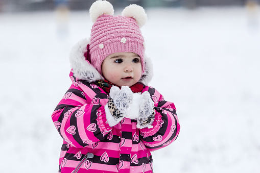 Portrait of little girl in pink jacket with red scarf and pink hat in snowy park at winter.