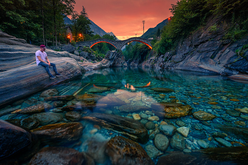 Tourist relaxes at the Verzasca river and enjoys colorful sunset above the double arch stone bridge at Ponte dei Salti in Lavertezzo, Canton Tessin, Switzerland