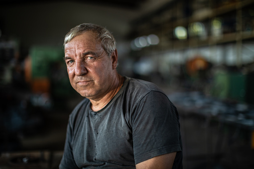 Portrait of mature manual worker in metal industry, sitting on table and looking at camera