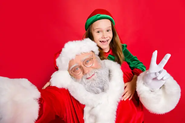 Close up photo of redhair elf  in green hat headwear and santa claus with white hair making selfie v-signs wearing eyewear eyeglasses isolated over red background