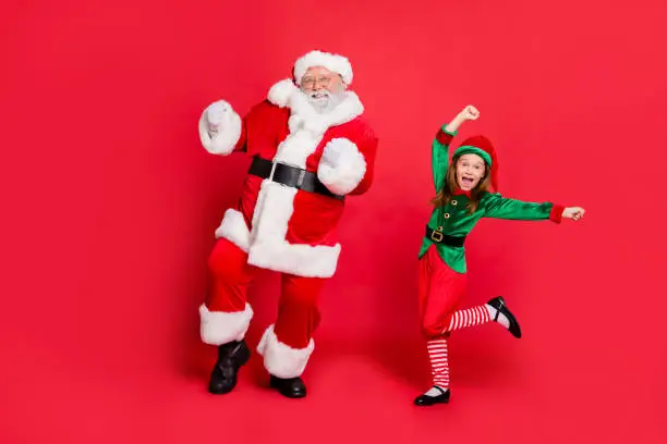 Full body photo of excited two santa claus in hat headwear dancing on festive event  wearing green bright costume isolated over red background