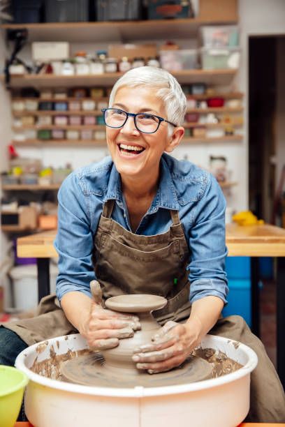 oleiro fêmea sênior que trabalha na roda da cerâmica ao sentar-se em sua oficina - potter human hand craftsperson molding - fotografias e filmes do acervo