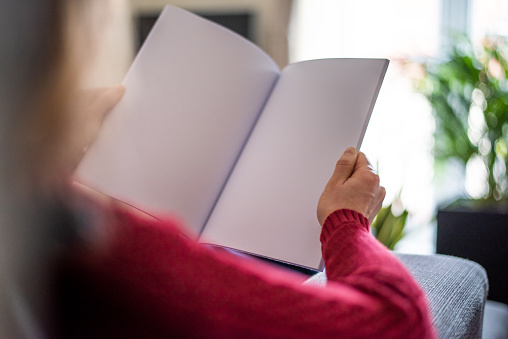 A woman is sitting on a couch in a living room and is holding a paper magazine in her hands. The publication is open and you can see two pages of the inside of the brochure. The pages are left blank for your own design ideas.