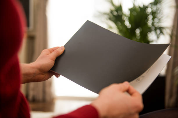 A woman is holding a paper magazine and is opening the front page of the publication. She is sitting in a living room at home while reading and looking at the paper document. The photo is taken out of a side view. The page is left blank for your own design template ideas.