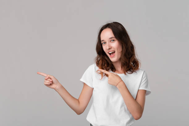 istock Pretty brunette woman in a white tee pointing at the empty space in a left side of a frame standing isolated over grey background. Copy space of advertising. 1177033447