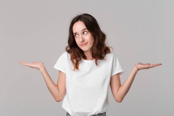 istock Pretty brunette woman in a white tee shrugging her shoulders beacause she doesn't know the answer standing isolated over grey background. Copy space of advertising. 1177033464