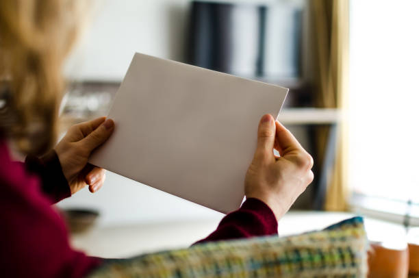 Woman reading blank magazine with copy space The woman is sitting on a sofa in a domestic living room and is holding a white brochure in her hands. The photo was taken out of a rear view perspective. The document is left empty for your own design ideas. over the shoulder view photos stock pictures, royalty-free photos & images