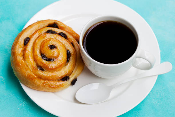 Fresh danish pastry with raisins with a cup of black coffee on blue table background. Fresh danish pastry with raisins with a cup of black coffee on blue table background black coffee swirl stock pictures, royalty-free photos & images
