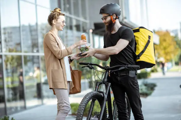 Courier delivering fresh lunches to a young business woman on a bicycle with thermal backpack. Takeaway restaurant food delivery concept