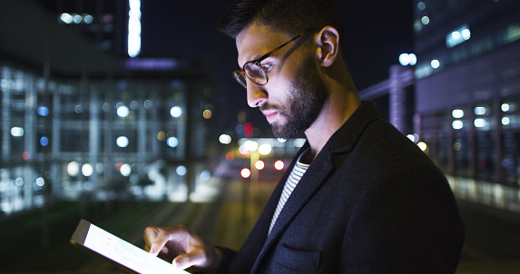 Cropped shot of a handsome young businessman standing alone and using a tablet in the office late at night