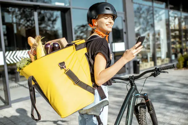 Young female courier delivering fresh food, standing with smartphone and backpack full of products outdoors
