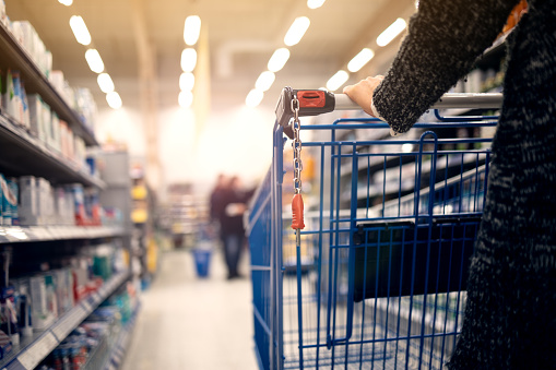 A women walks with a shopping basket in a store. Hand and part of the basket in focus, blurred background
