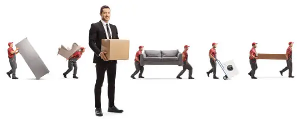 Photo of Man in a suit with a cardboard box and workers from a moving company carrying home appliences and furniture