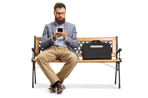 Bearded man typing on a mobile phone and sitting on a bench isolated on white background