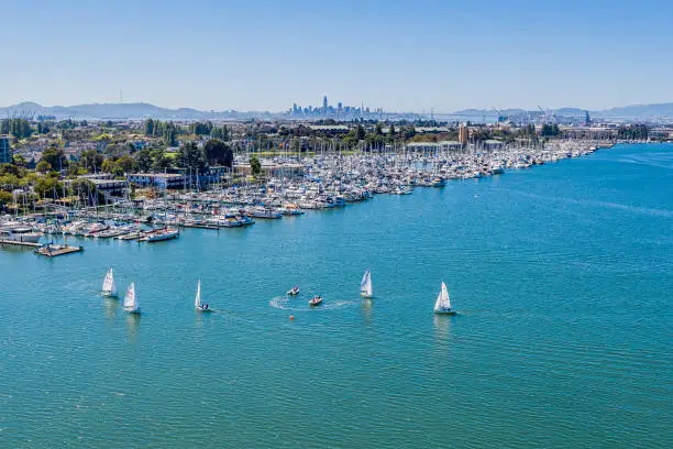 An aerial view of Alameda, Alameda Point , the skyline of San Francisco across the Bay and sailboats in the bay. A beautiful blue sky on a sunny day. The harbor is bustling with activity.