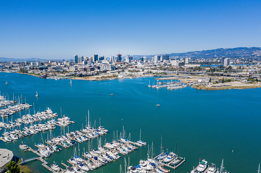 An aerial view of Alameda, Oakland skyline and the marina on a beautiful blue sky sunny day. The harbor is bustling with activity. The skyscrapers of Oakland are seen across the Bay.