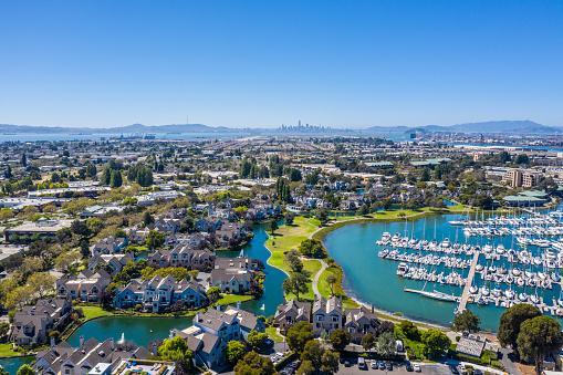 An aerial view of Alameda, Alameda Point , the skyline of San Francisco across the Bay and sailboats in the marina. A beautiful blue sky on a sunny day. Luxury homes along the shoreline.