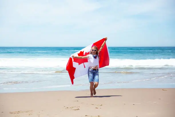 Happy Canadian girl carries fluttering white red flag of Canada against blue sky and ocean background. Canadian flag is a symbol of freedom, liberty, democracy, independence. Canada Day