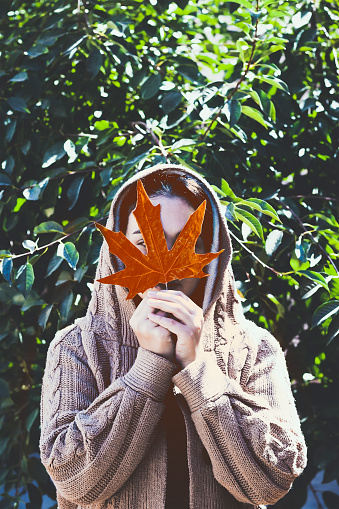 Fall season concept. A beautiful middle-eastern woman holds a yellow autumn leaf. Young female hiding her face with fall leaves. Autumn or fall woman. Sunny autumn weather at outdoors.