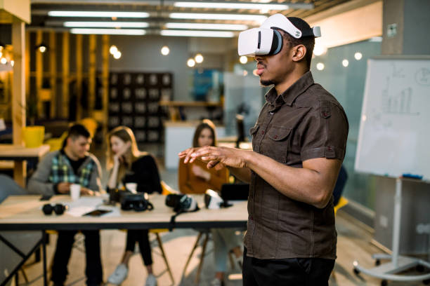 istock African young man wearing virtual reality glasses in modern interior design coworking studio. Smartphone using with VR goggles headset. Team of young people working at the table on the background 1177053038
