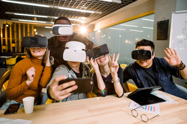 istock Five young funny people sitting at the table in front of each other, using virtual reality goggles, having fun and gesturing. VR goggles concept, business meeting. 1177055209