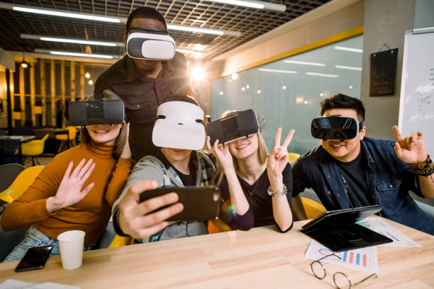 istock Five young funny people sitting at the table in front of each other, using virtual reality goggles, having fun and gesturing. VR goggles concept, business meeting. 1177055319